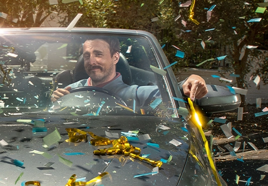 Ein freundlicher Mann sitzt stolz in einem Auto, es regnet goldenes Konfetti.
